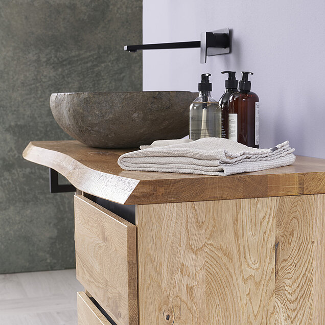 Waschtisch mit einer hölzernen Arbeitsplatte, einem steinernen Waschbecken und einem an der Wand montierten schwarzen Wasserhahn.