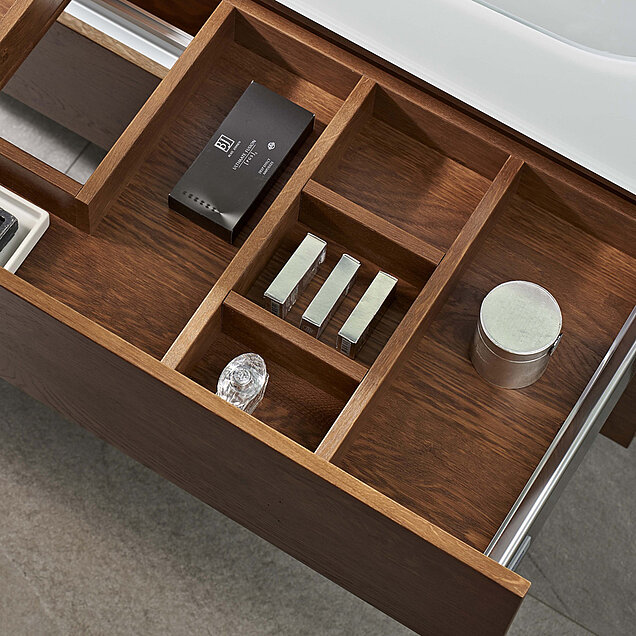 Offene Holzschublade mit Fächern mit drei kleinen rechteckige Flaschen und einen runden silbernen Behälter neben einem Waschbecken.