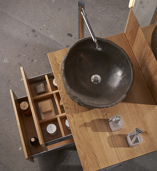 Dunkles Steinwaschbecken auf einer hölzernen Arbeitsplatte. Darunter befindet sich eine offene Schublade mit Toilettenartikeln.