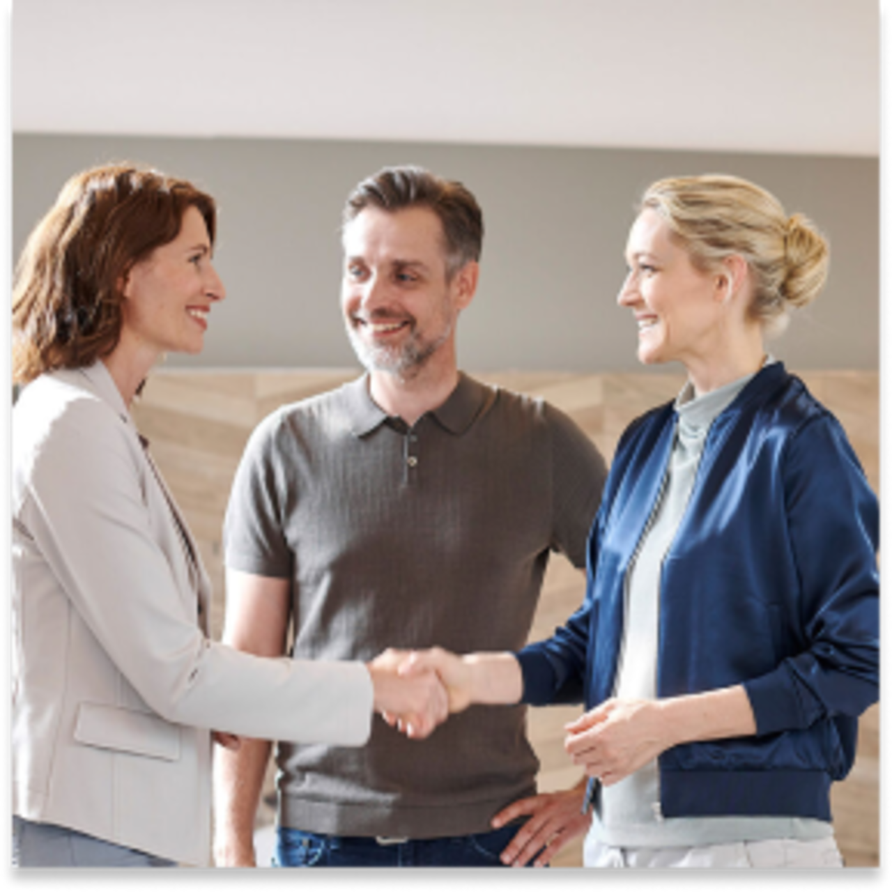 Zwei Frauen schütteln sich die Hände und lächeln, während ein Mann neben ihnen steht und ebenfalls lächelt. 