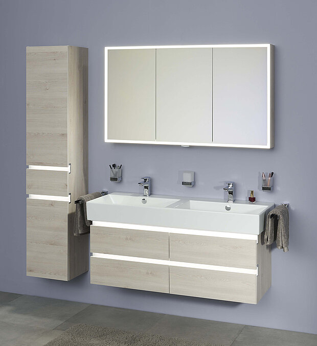 Badezimmer mit Waschtisch aus hellem Holz, einem Doppelwaschbecken, zwei Wasserhähnen, einem hohen Schrank und einem beleuchteten Spiegel.