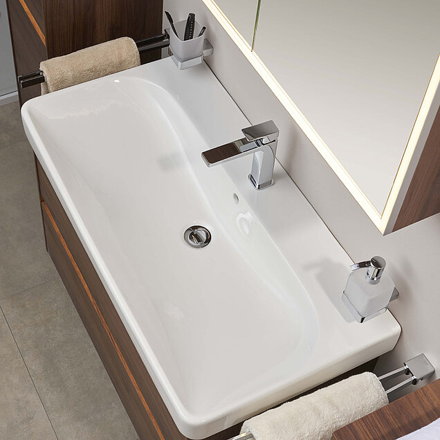 Weißes, rechteckiges Waschbecken mit verchromtem Wasserhahn, Seifenspender und Zahnbürstenhalter, unter einem großen Spiegel.