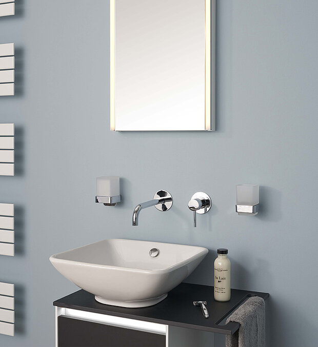 Bad mit rechteckigem Spiegel, Wandarmatur und Seifenspendern über einem quadratischen weißen Waschbecken.