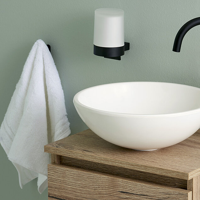 Badezimmer mit einem weißen Handtuch an einem schwarzen Haken, einem weißen Waschbecken auf einem hölzernen Waschtisch, einem schwarzen Wasserhahn und einem an der Wand befestigten Seifenspender