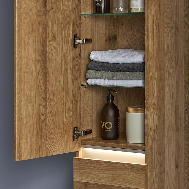 Ein offener hölzerner Badezimmerschrank mit Glasfächern für gefaltete Handtücher und Toilettenartikel.