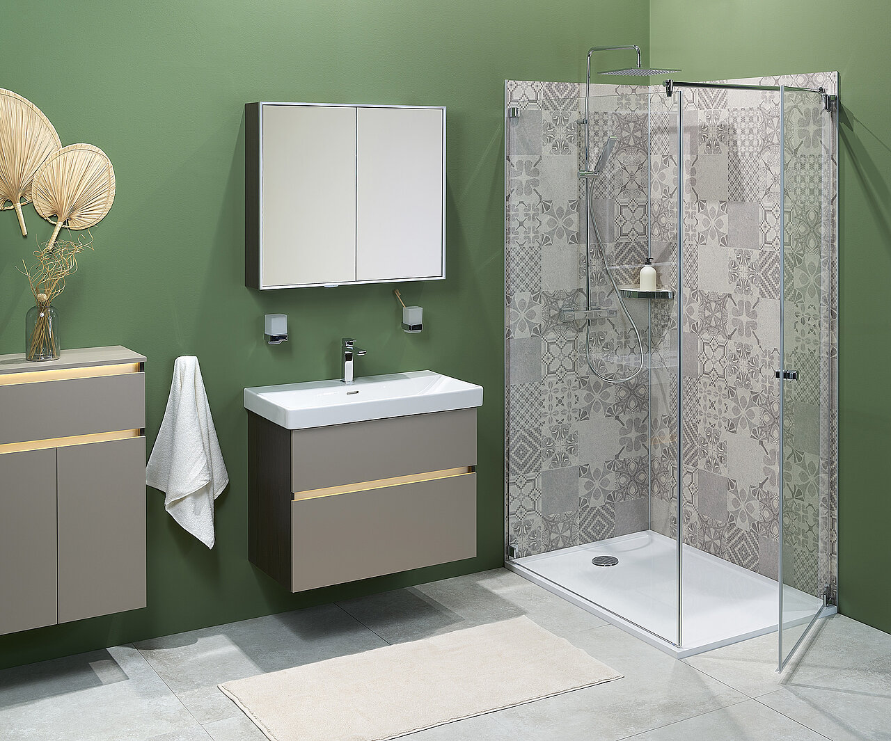 Badezimmer mit grünen Wänden, Waschtisch und Waschbecken, Spiegelschrank und einer Dusche mit Glasabtrennung und gemusterter Duschrückwand. 