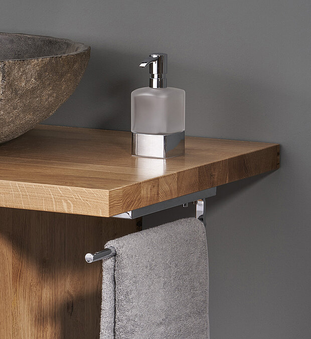 Eine moderne Badezimmer-Arbeitsplatte aus Holz mit einem Waschbecken aus Stein, einem Seifenspender aus Milchglas und einem grauen Handtuch.