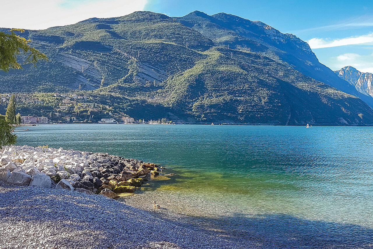 Der klarer blauer Gardasee mit felsigem und kieseligem Ufer, mit grünen, steil aufragenden Bergen im Hintergrund und blauen Himmel.