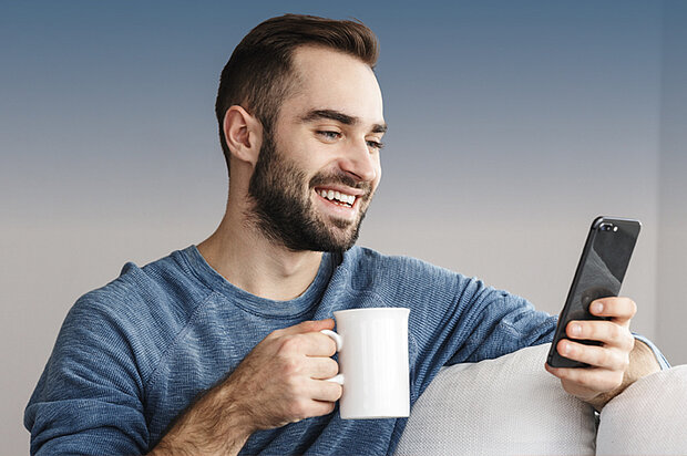 Ein lächelnder Mann mit Bart, der einen blauen Pullover trägt, sitzt auf einer Couch und hält in der einen Hand einen weißen Becher, während er in der anderen auf sein Smartphone schaut.