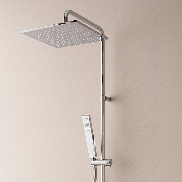 Ein chromfarbener eckiger Brausekopf einer Dusche und eine beige Wand im Hintergrund