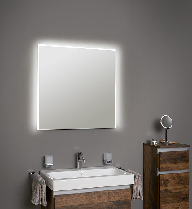 Badezimmer mit einem großen beleuchteten quadratischen Spiegel, einem weißen Waschbecken mit verchromtem Wasserhahn.