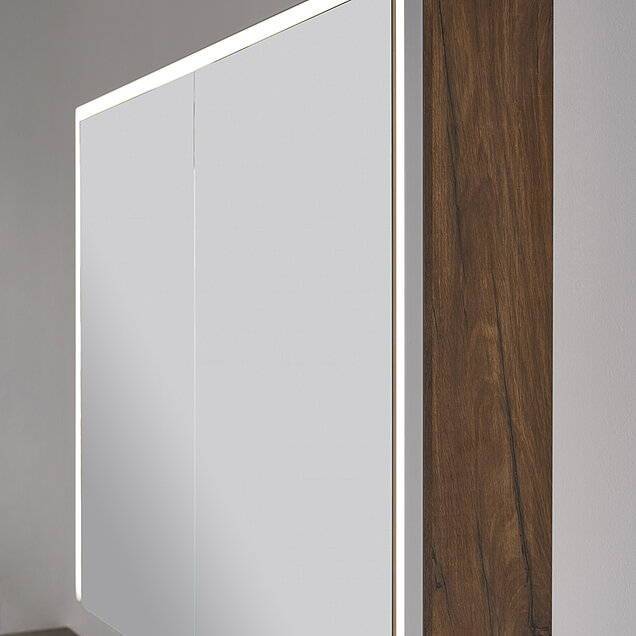 Eine Nahaufnahme eines modernen Badezimmerschranks mit Spiegeltüren und einer Seitenwand aus Holz. Der Spiegel verfügt über eine integrierte LED-Lichtleiste 