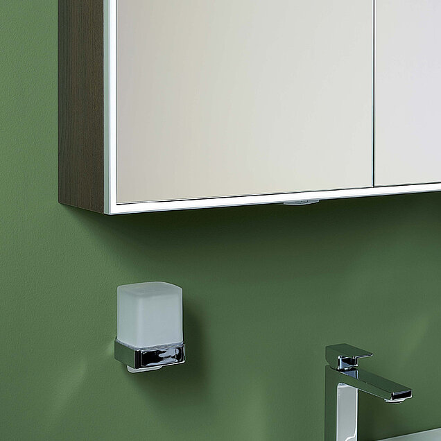 Ein an der Wand montierter Spiegelschrank, ein quadratischen Seifenspender aus Milchglas mit Chromhalterung und ein verchromter Wasserhahn.