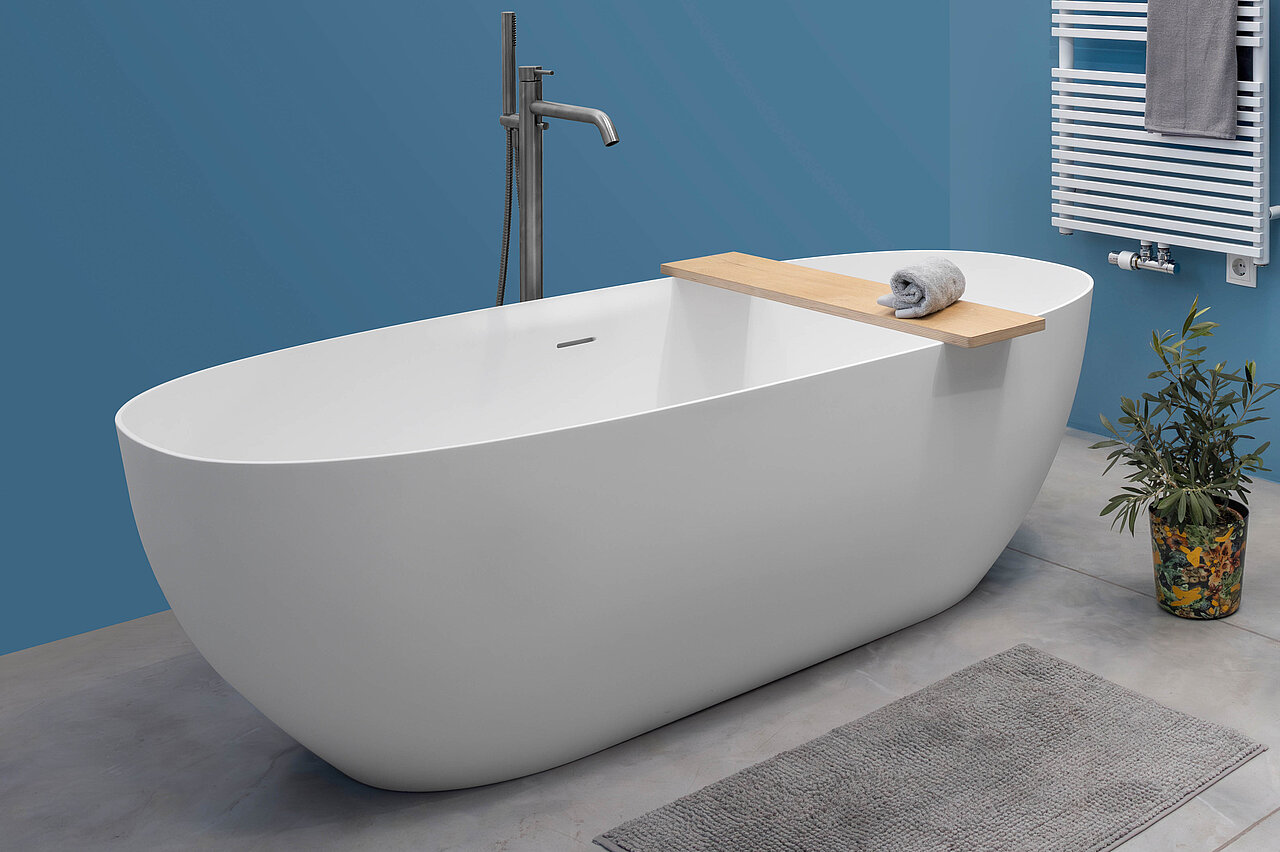 Neue Larimar CAPRI Badewanne mit modernen Rundungen und grauer samtweichem Oberflächenmaterial names SolidSurface ist freistehend im Raum montiert mit einer matten chromfarbenen Standarmatur in einem blau gestrichenen Badezimmer welches mit schwarzen Badmöblen einem Siegelschrank und einer Handtuchheizung kombiniert ist.