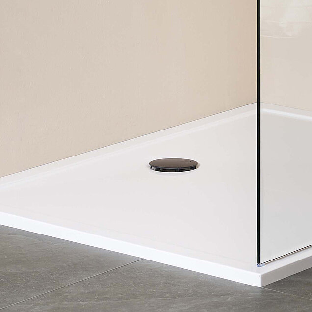 Ein moderner, minimalistischer Duschbereich mit einer weißen Duschwanne, einem runden schwarzen Ablauf, einer Glaswand und grauen Bodenfliesen. 