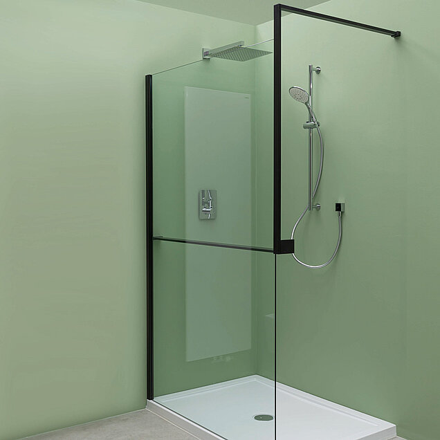 Minimalistische Dusche mit transparenten Glaswänden, schwarzem Metallrahmen, Kopf- und Handbrausen und einem hellgrünen Wandhintergrund.