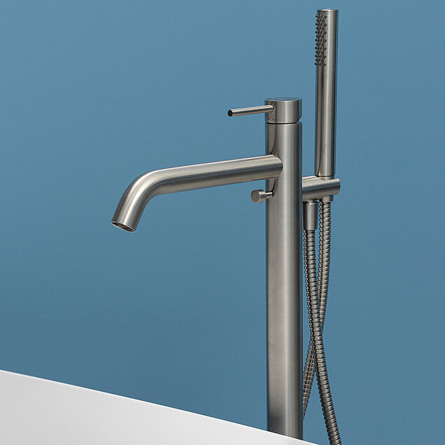 Eine moderne Edelstahl-Badewannenarmatur mit seitlichem Griff und aufgesetzter Handbrause vor einer schlichten hellblauen Wand. 