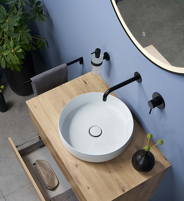 Ein modernes rundes Waschbecken auf einem hellen Holzwaschtisch, schwarzem Wasserhahn und Armaturen und einem Spiegel.