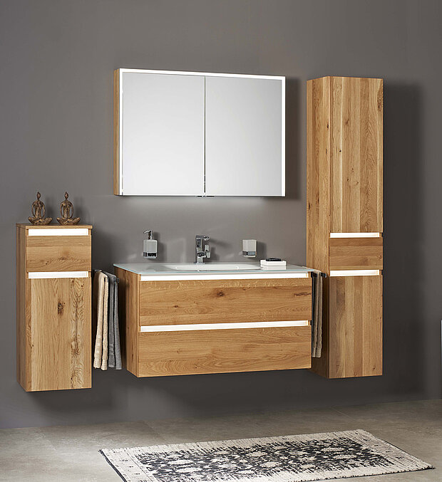 Badezimmer mit einem großen beleuchteten Spiegel, schwebenden Holzschränken und einem Waschbecken mit Chromarmaturen. 