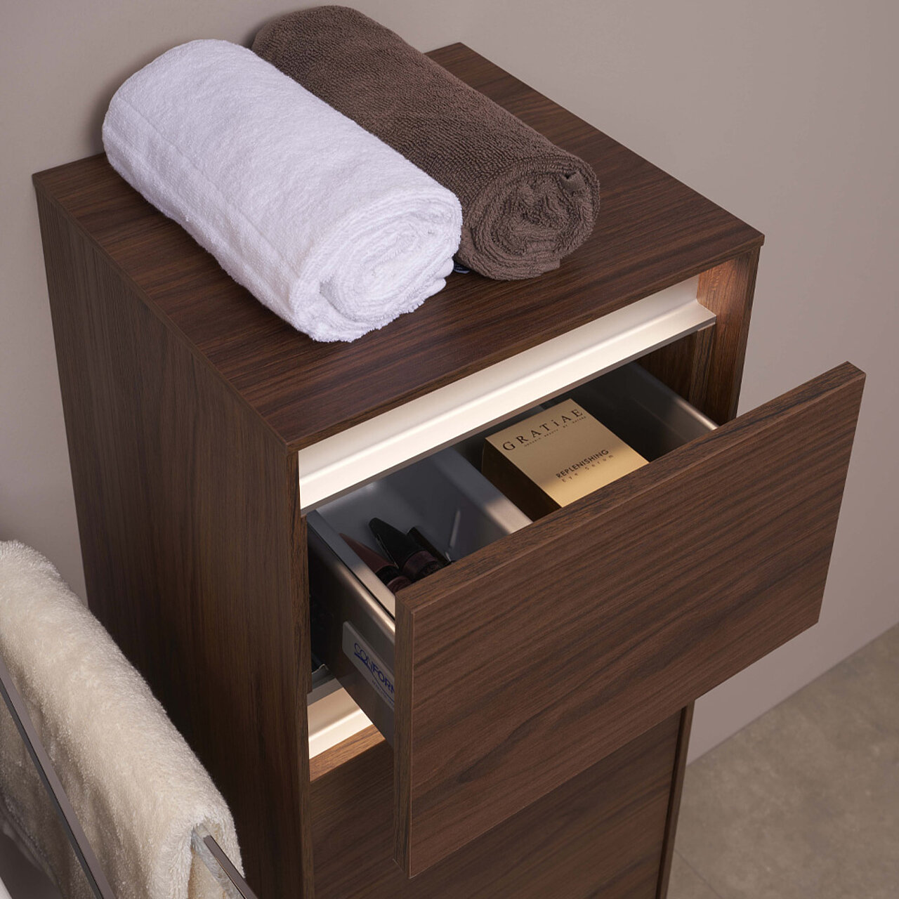 Holzschrank mit einer offenen Schublade, in der sich eKosmetika befinden; darauf liegen zwei gerollte Handtücher
