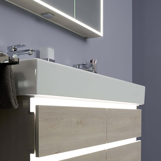 Rechteckiges weißes Waschbecken mit hellem Holzschrank, LED-Beleuchtung und einem seitlich hängenden Handtuch. 