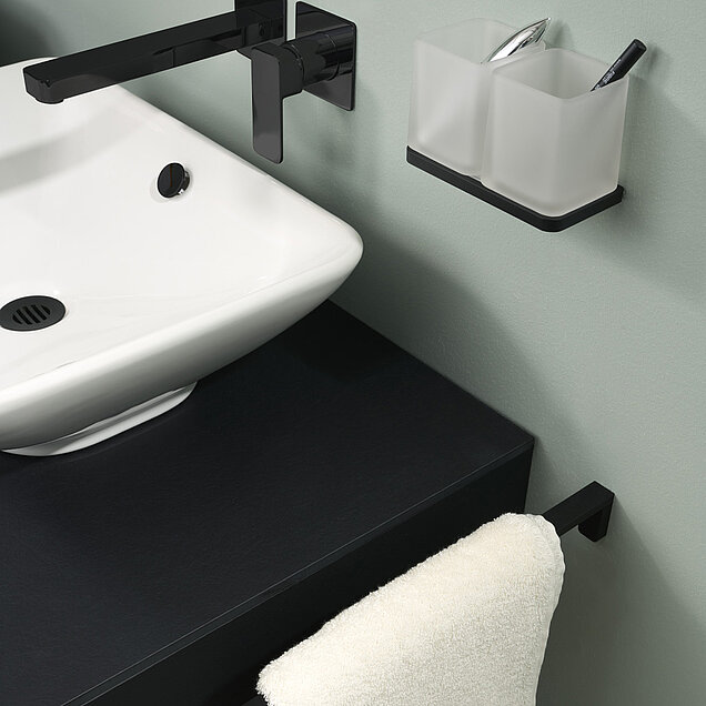 Ein Waschbecken mit schwarzen Wasserhahn, einer schwarzen Arbeitsplatte, einem weißen Handtuch auf einem schwarzen Handtuchhalter und zwei mattierten Bechern an der Wand.