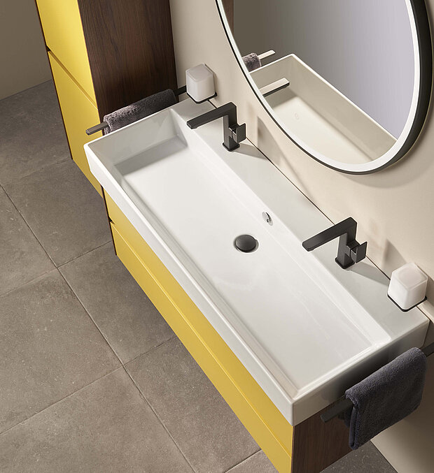 Weißes rechteckiges Waschbecken mit zwei mattschwarzen Wasserhähnen, gelben Schränken und einem runden Spiegel.