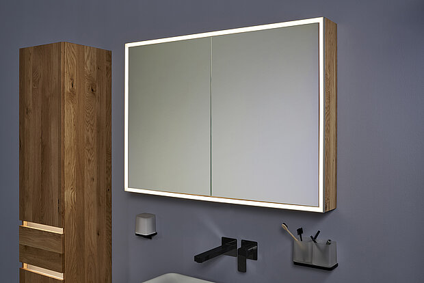 Ein modernes Badezimmer verfügt über einen Holzschrank mit Schubladen und einen Wandspiegelschrank mit integrierter Beleuchtung.