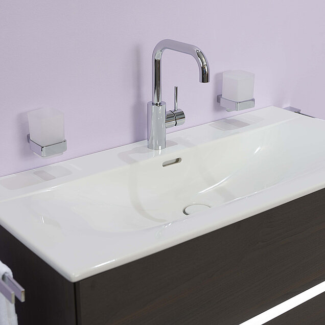 Weißes Waschbecken mit einem verchromten Wasserhahn, montiert auf einem Waschtisch aus dunklem Holz vor einer hellen Wand.