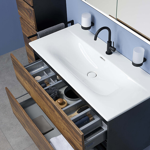 Waschbecken mit schwarzem Wasserhahn, zwei an der Wand befestigte Becherhalter und offene Holzschubladen mit Toilettenartikeln.