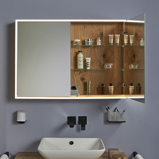 Ein Badezimmer-Spiegelschrank, teilweise geöffnet, um Regale mit Toilettenartikeln, Cremes und Flaschen freizulegen. 